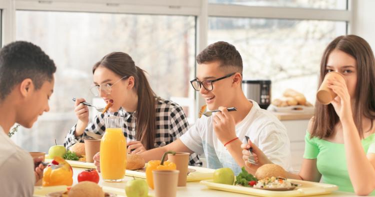 Teenagers enjoying a bright and vibrant lunchtime together, sharing smiles and conversations over a table filled with fresh fruit, sandwiches, and juice in a sunny cafeteria setting.